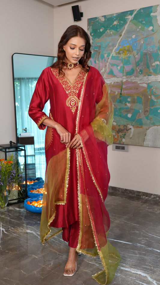 A woman models a traditional Indian outfit consisting of a red kurta with gold embroidery, a matching red skirt, and a sheer gold dupatta draped over her shoulders, set in an indoor room with a large abstract painting on the wall.